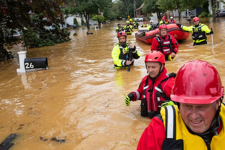 Uma fila de equipes de resgate em coletes brilhantes e capacetes caminha com água até a cintura em uma rua inundada, puxando uma jangada. A água está na caixa de correio que eles estão passando.