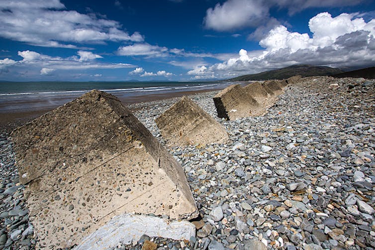 Uma série de cubos de concreto se alinham em uma praia de seixos.
