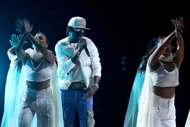 A man in a padded waistcoat with a cap and sunglasses on performs into a mic as women in white with flowing fabric dance around him, their hands crossed in the air in front of them.
