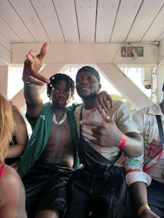 Two young African men sit, each with one arm around the other man and the other arm making hand gestures to camera.