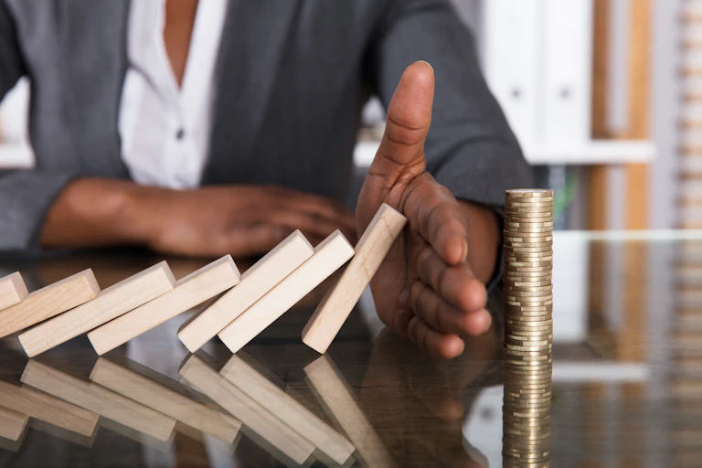 Close-up Of A Human Hand Stopping Wooden Blocks From Falling On Stacked Coins Over Desk