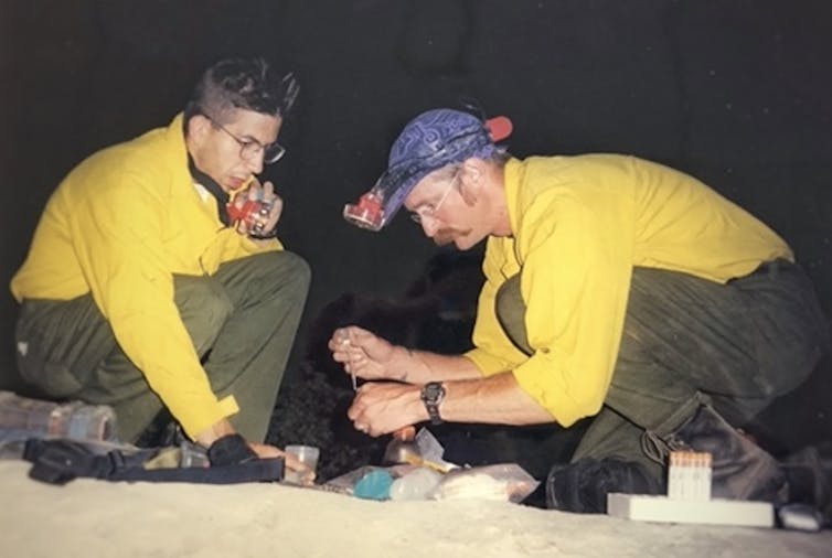 A man wearing a head lamp leans over a set of vials with an eyedropper, while a firefighter in a yellow jacket sits nearby.