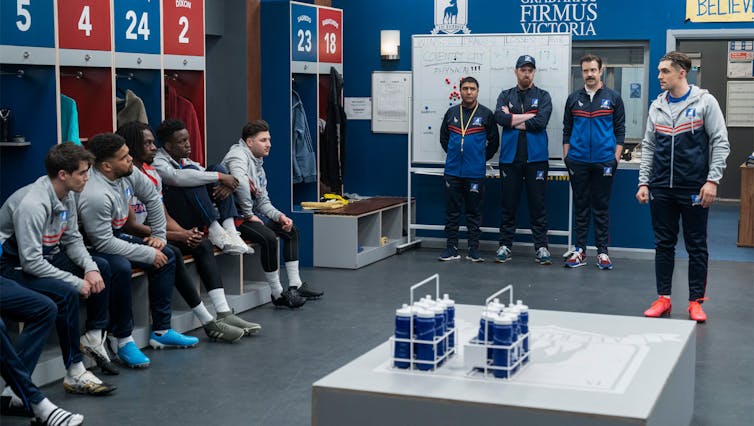 Ted Lasso and his AFC Richmond players in the dressing room. Ted and the other coaches stand in front of a whiteboard filled with tactics.