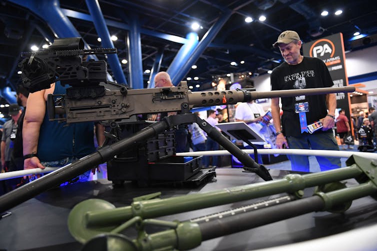 White men look at a machine gun on display in a crowded room with high ceilings