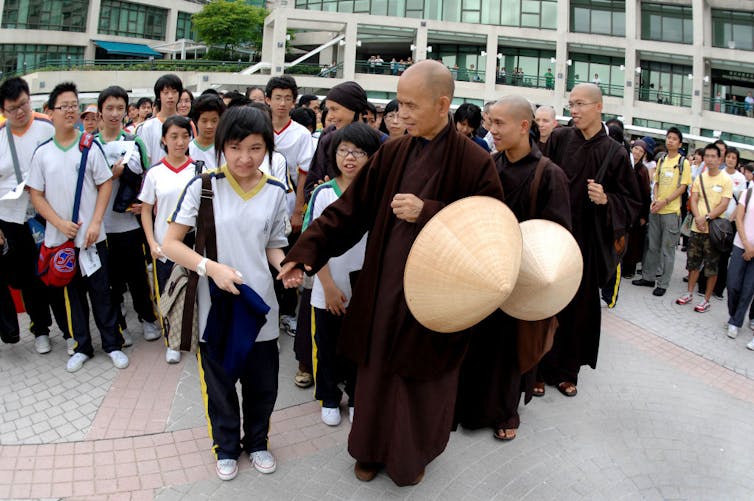 A row of monks stand next to a small crowd of schoolchildren in uniform as one monk takes a child's hand.