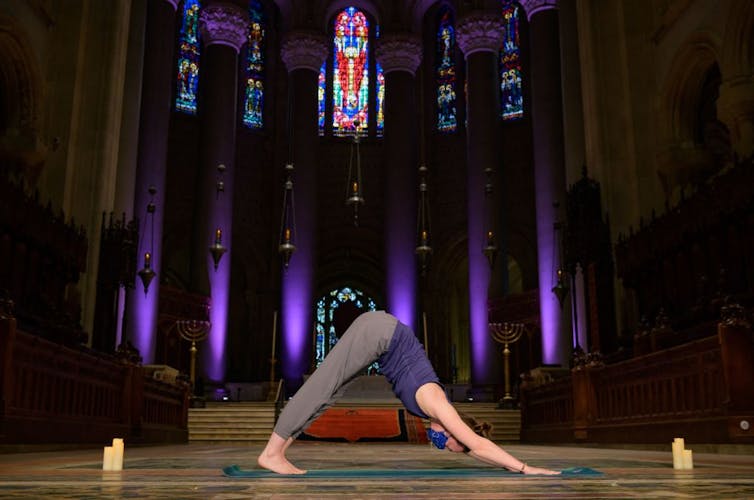 A woman in exercise clothes does a yoga pose inside a dark cathedral with stained glass windows.