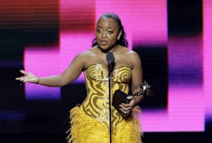 A woman wearing a yellow dress stands at the microphone holding a trophy.