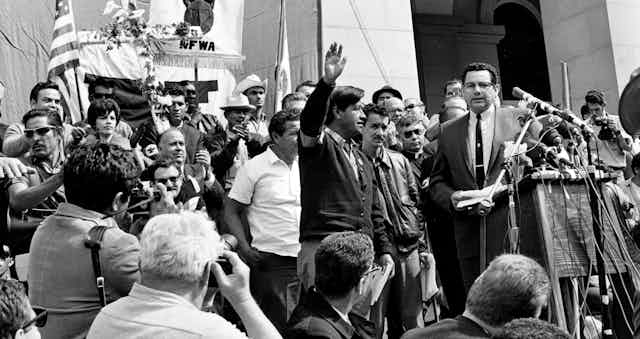 A man stand with his hand raised surrounded by a group in front of microphones.