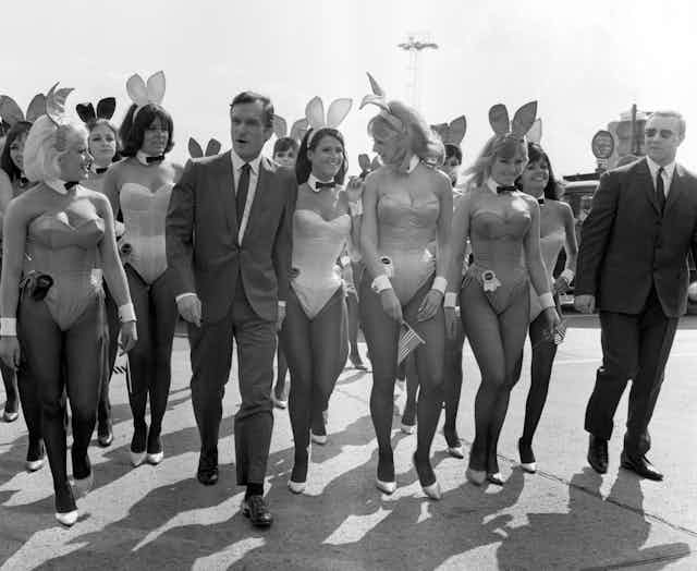 Black and white photograph of Hugh Hefner in a suit flanked by women in Playboy bunny costumes. 
