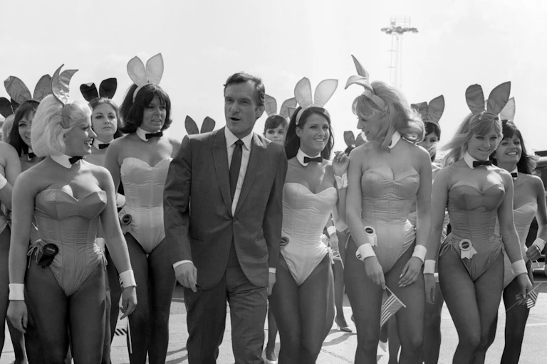 Black and white photograph of Hugh Hefner in a suit flanked by women in Playboy bunny costumes.
