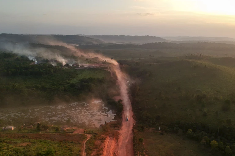 Smoke from fires rises near a road with a vehicle on it and a vast landscape behind it