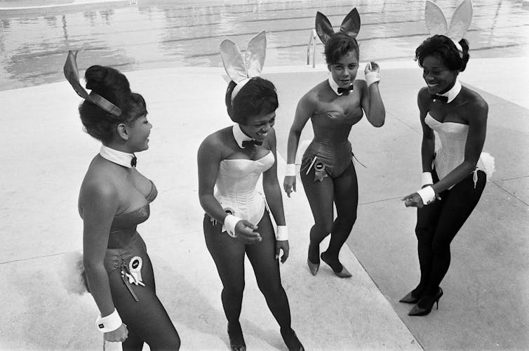 Four young black women in Playboy 'bunny' outfits dance and laugh in front of the camera.