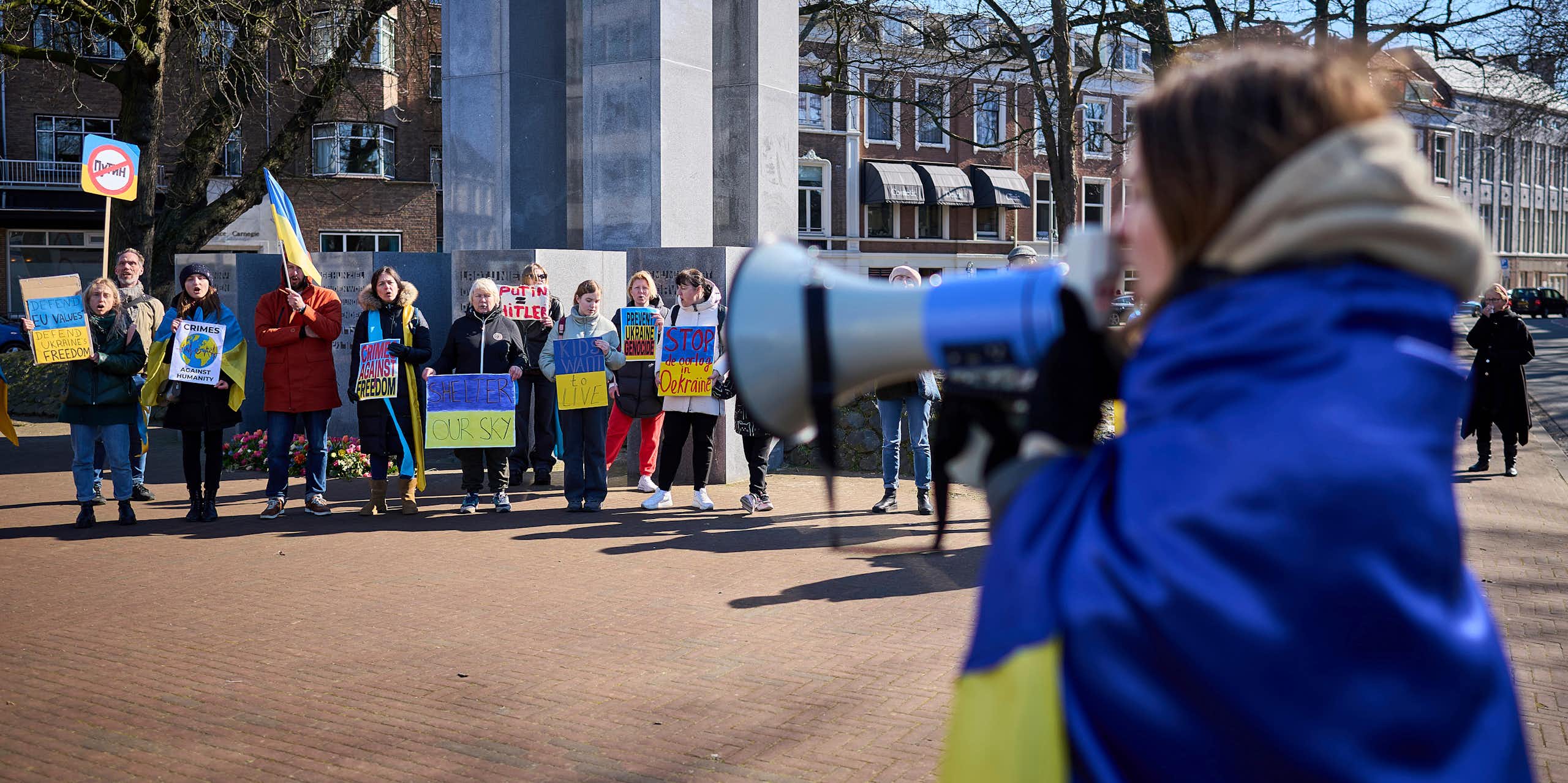 Une femme enveloppée dans un drapeau bleu et jaune crie dans un mégaphone. D'autres personnes tenant des panneaux bleus et jaunes se tiennent devant elle.