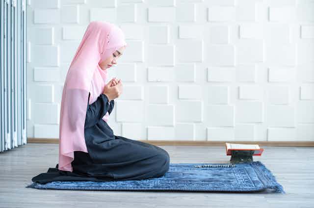 A young girl wearing a pink headscarf kneels on a blue prayer rug. The Qur'an is open on a small stand nearby.