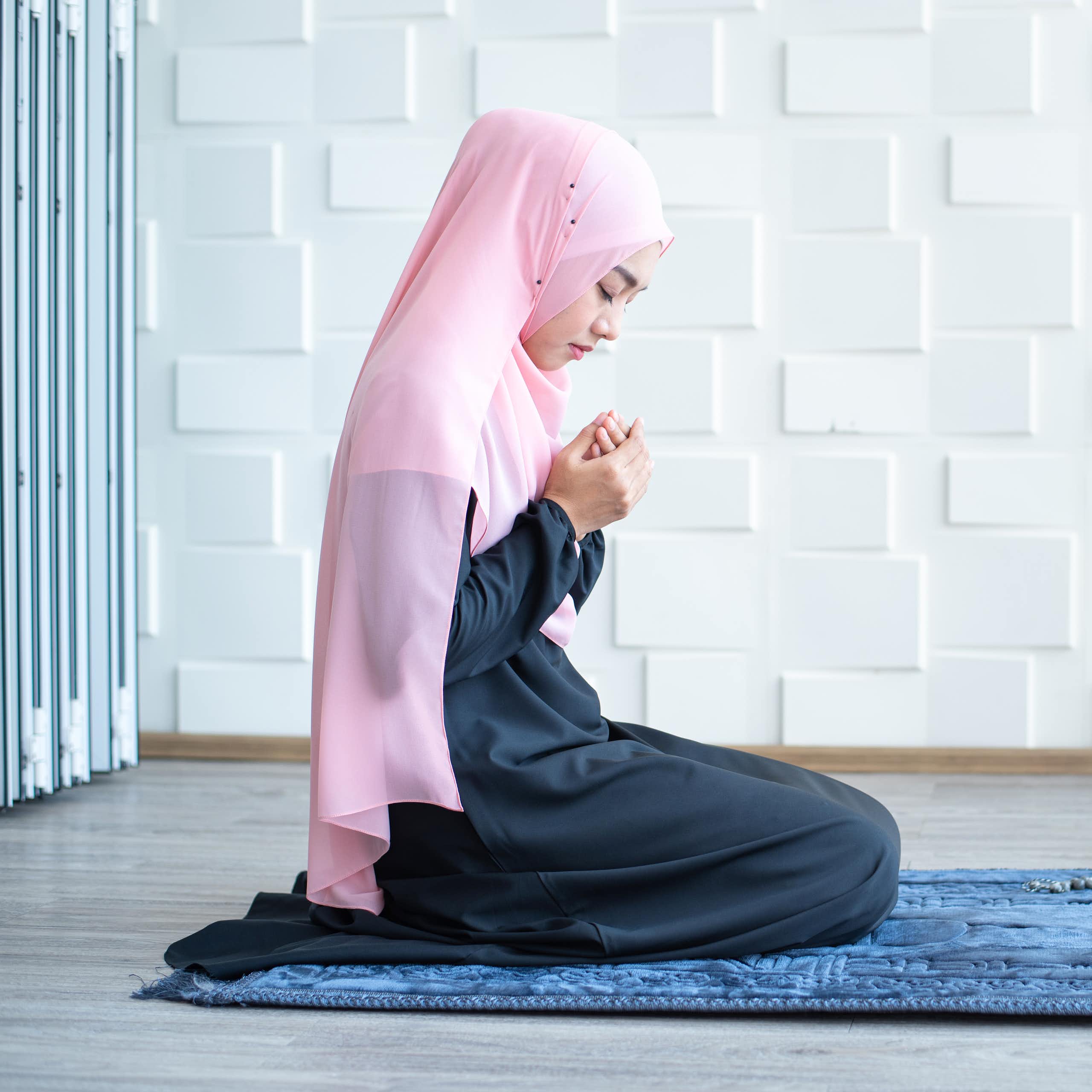 A young girl wearing a pink headscarf kneels on a blue prayer rug. The Qur'an is open on a small stand nearby.