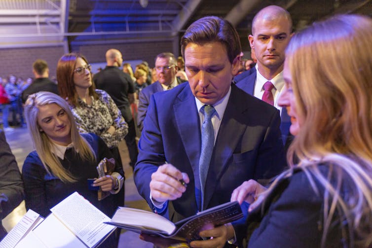 Con un grupo de personas de pie cerca, Ron DeSantis firma una copia de su libro.