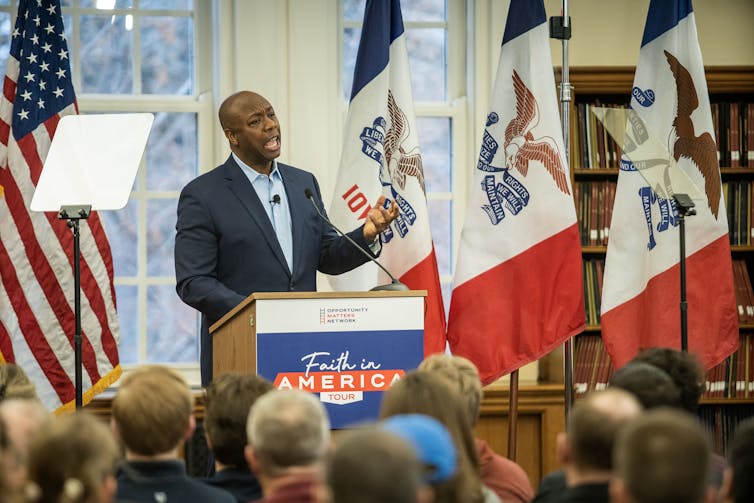 Standing in front of the U.S. and Iowa flags, Tim Scott speaks before the podium to a large crowd.