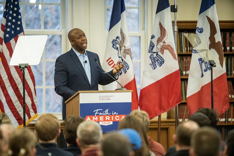 De pie frente a las banderas de EE. UU. e Iowa, Tim Scott habla ante una gran multitud ante el podio.