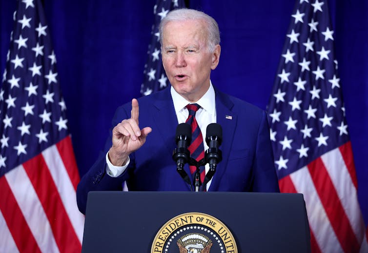 President Biden, in dark blue jacket, red and blue striped tie, and white shirt, speaks at a podium adorned with the Presidential Seal in front of two American flags.