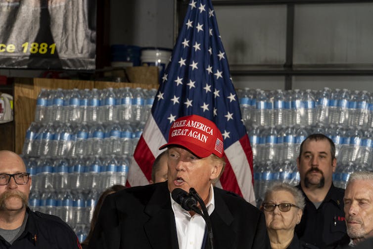 El expresidente Trump, con un sombrero MAGA y parado frente a una paleta de agua embotellada, habla a una multitud de simpatizantes.