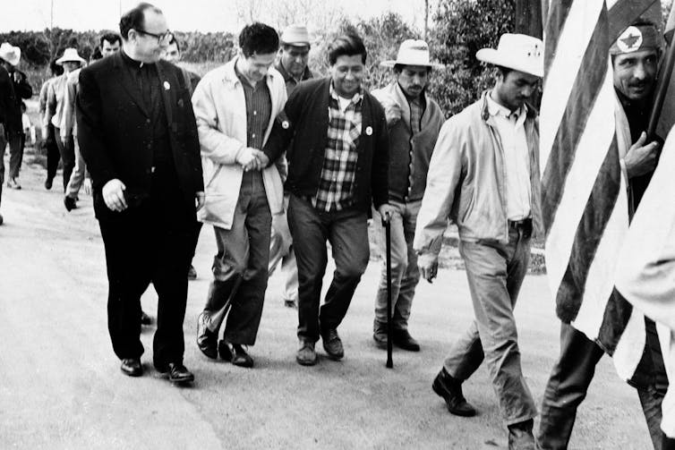 A man in seen walking while leaning on a cane, surrounded by a group of supporters including a man in vicar garb.