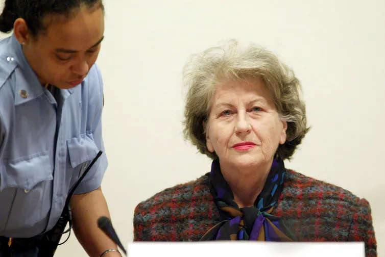 Biljana Plavsic sits at her trial at the Hague, with a female UN guard leaning over her