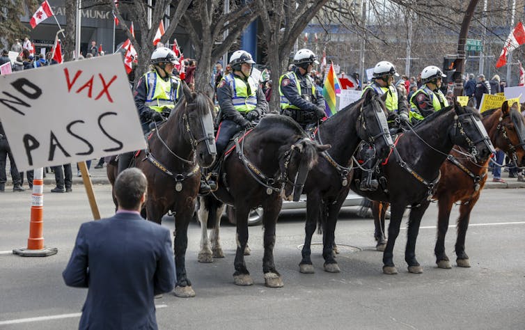 A row of police officers on horseback stand in front of a man carrying a sign reading No Vax Pass.
