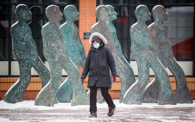 A woman in a winter coat and hood wears a mask while walking past an art installation of people walking on a city street.