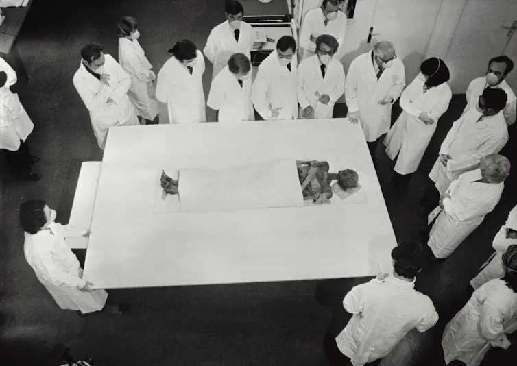 Photo d’époque, vue en plongée, de la momie entourée de savants en blouse blanche