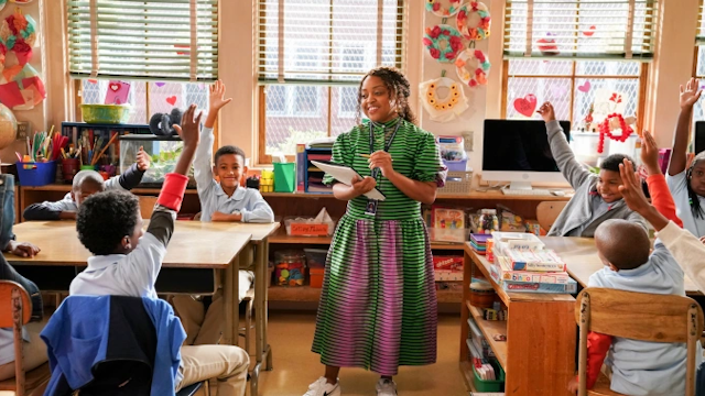 A teacher in a black and green stripped dress with hints of pink, stands before a classroom of elementary school students seated at desks with their hands raised.