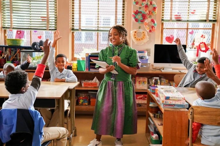 A teacher in a black and green stripped dress with hints of pink, stands before a classroom of elementary school students seated at desks with their hands raised.