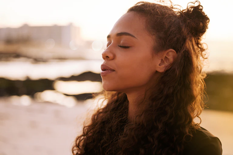 A woman with dark curly hair breathes deeply with her eyes closed near the ocean.