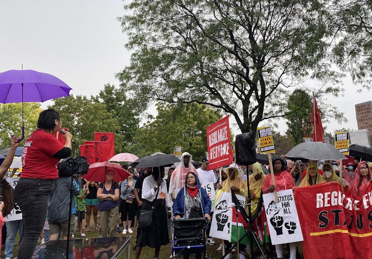 A group of people at a protest carrying signs that read: status for all. A woman in a red shirt speaks into a mic.