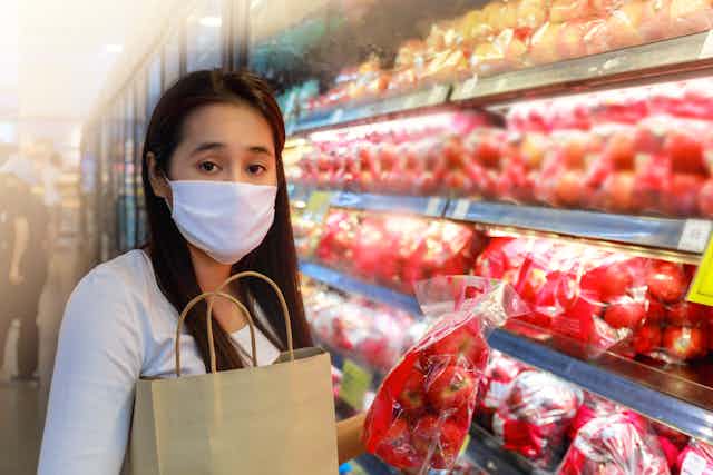 Asian woman wearing protective face mask holding bag of apples in supermarket produce aisle. 