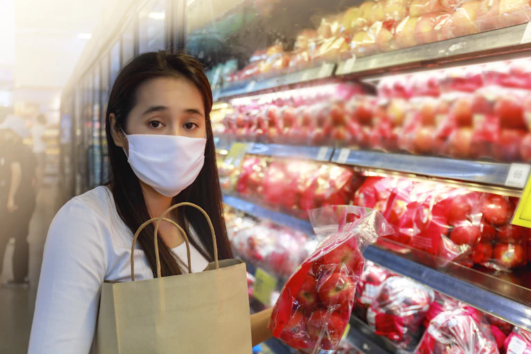 Asian woman wearing protective face mask holding bag of apples in supermarket produce aisle.
