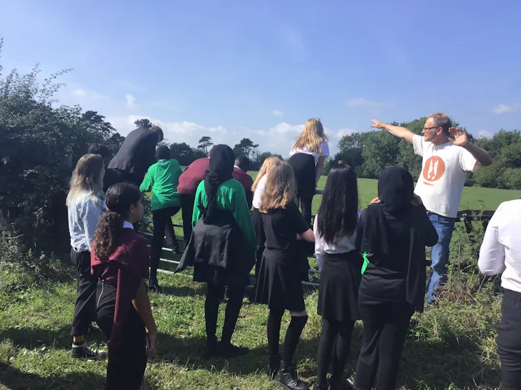 A group of school children stand in a field with their backs towards the camera. A man stands in front of them pointing towards something.