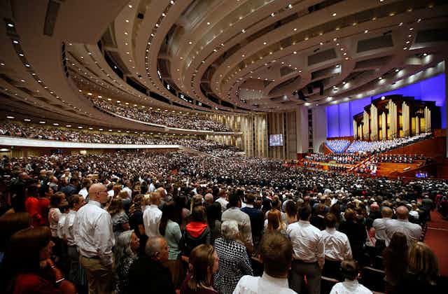 A large crowd of people in a fancy auditorium stand looking at a choir in the front.
