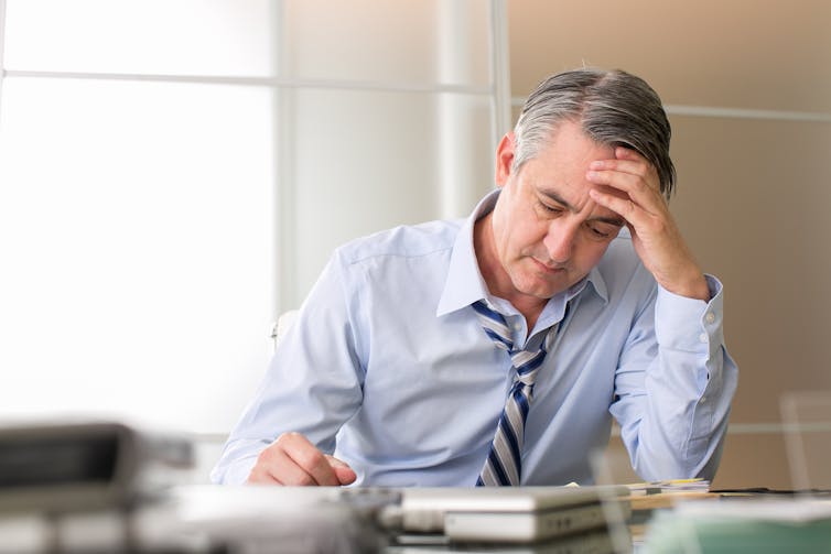 A middle-aged man, with a frustrated look on his face, leans his head against his hand as he looks at paperwork on his desk