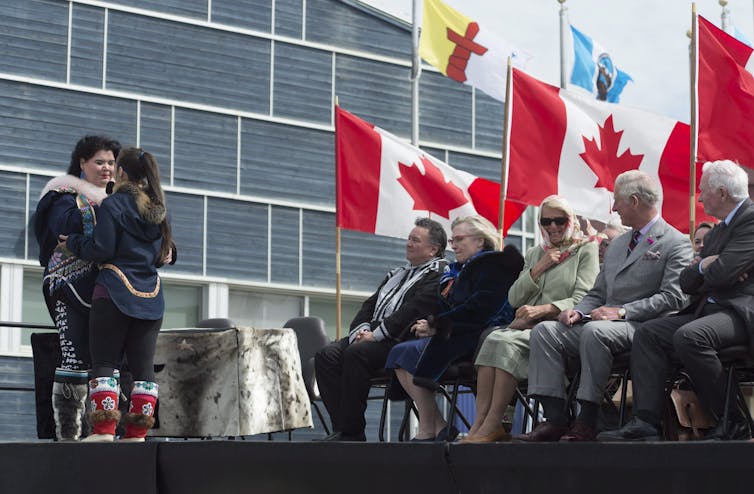 Two women stand to one side while seated people watch them. Canadian and Nunavut flags fly in the background.