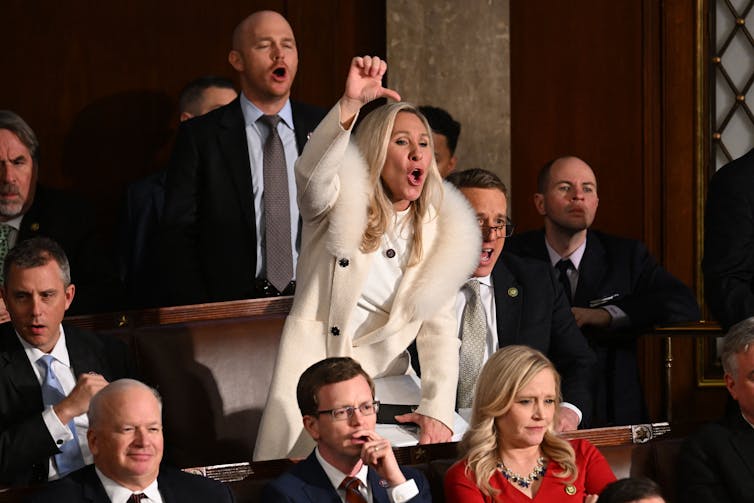 A standing woman, in a white coat with fur around the collar, makes a thumbs down gesture as seated people surrounded her.