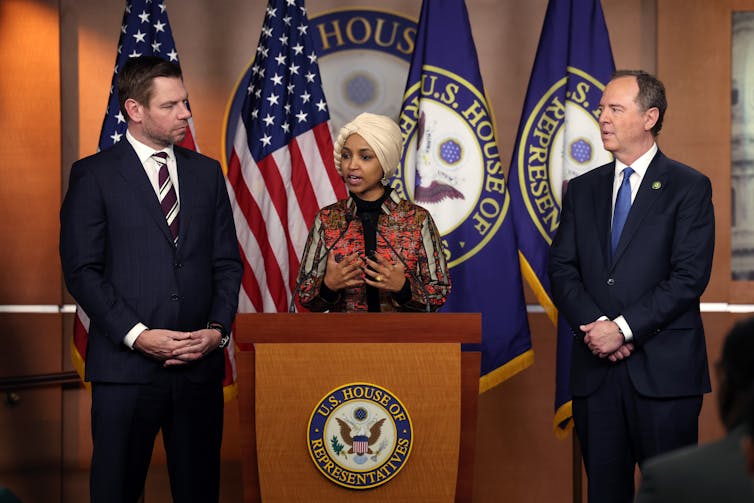 Standing before a podium, a woman speaks as two men, with their hands clasped, watch.