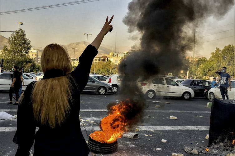 A woman face away from the camera stands in front of a burning tire and raises her hand and makes a victory sign.