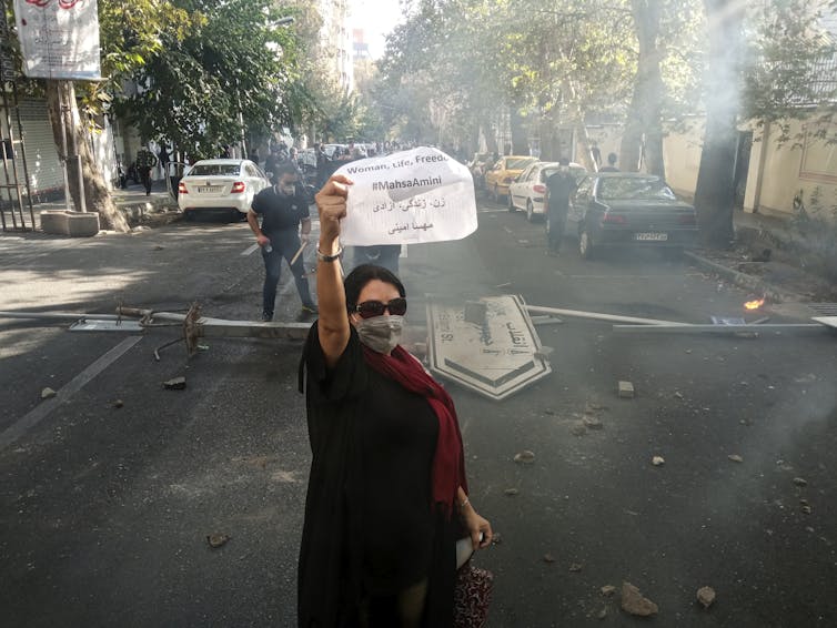A woman in a face mask and sunglasses carries a sign that reads: women life freedom