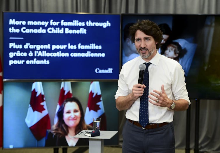 A man in a dress shirt and tie speaks into a microphone while a smiling woman is displayed on a large screen behind him