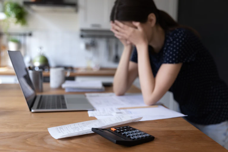 A young woman leans against a counter with her face in her hands. On the counter in front of her is a pile of papers, an open laptop and a calculator.
