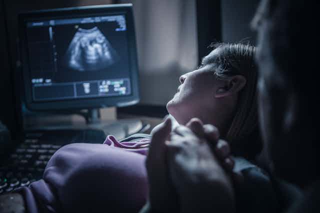 A pregnant couple in an exam room look over at a sonogram screen.