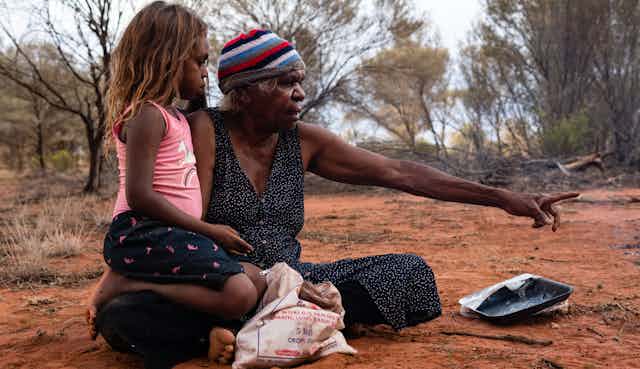 First Nations grandmother sitting on ground with young girl, pointing into the bush