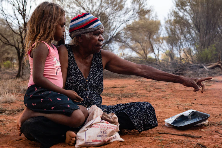 First Nations grandmother sitting on ground with young girl, pointing into the bush