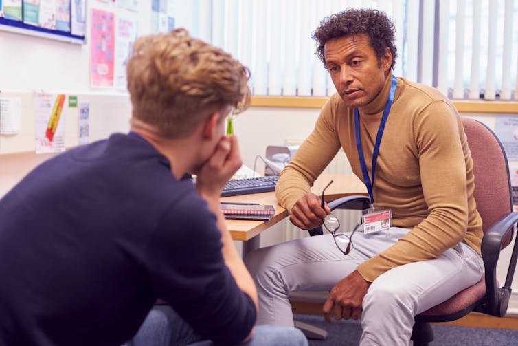 A teacher speaks to a teenage student in an office.