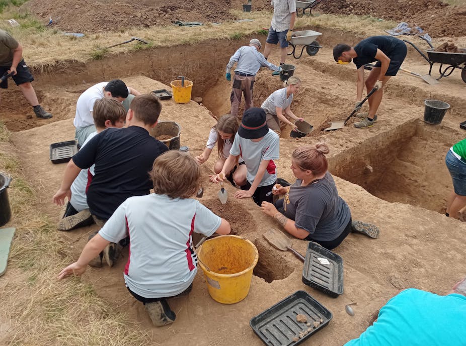 A group of children and adults gather around various excavated holes in the ground with buckets and shovels. 